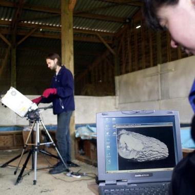 A laptop screen displaying a laser scan of one of the Seahenge timbers. In the background, a man is using a laser scanner on a tripod to scan the timber.