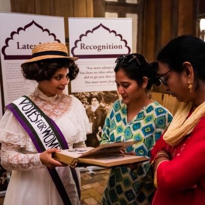 Actress dressed as Sophia Duleep Singh showing a book to two museum visitors