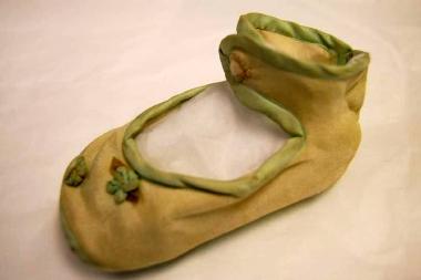 Yellow fabric infants shoe from the 1940s. The shoe has an ankle strap, the edges are bound with green silk ribbon and there are two small ribbon flowers on the front of the shoe.
